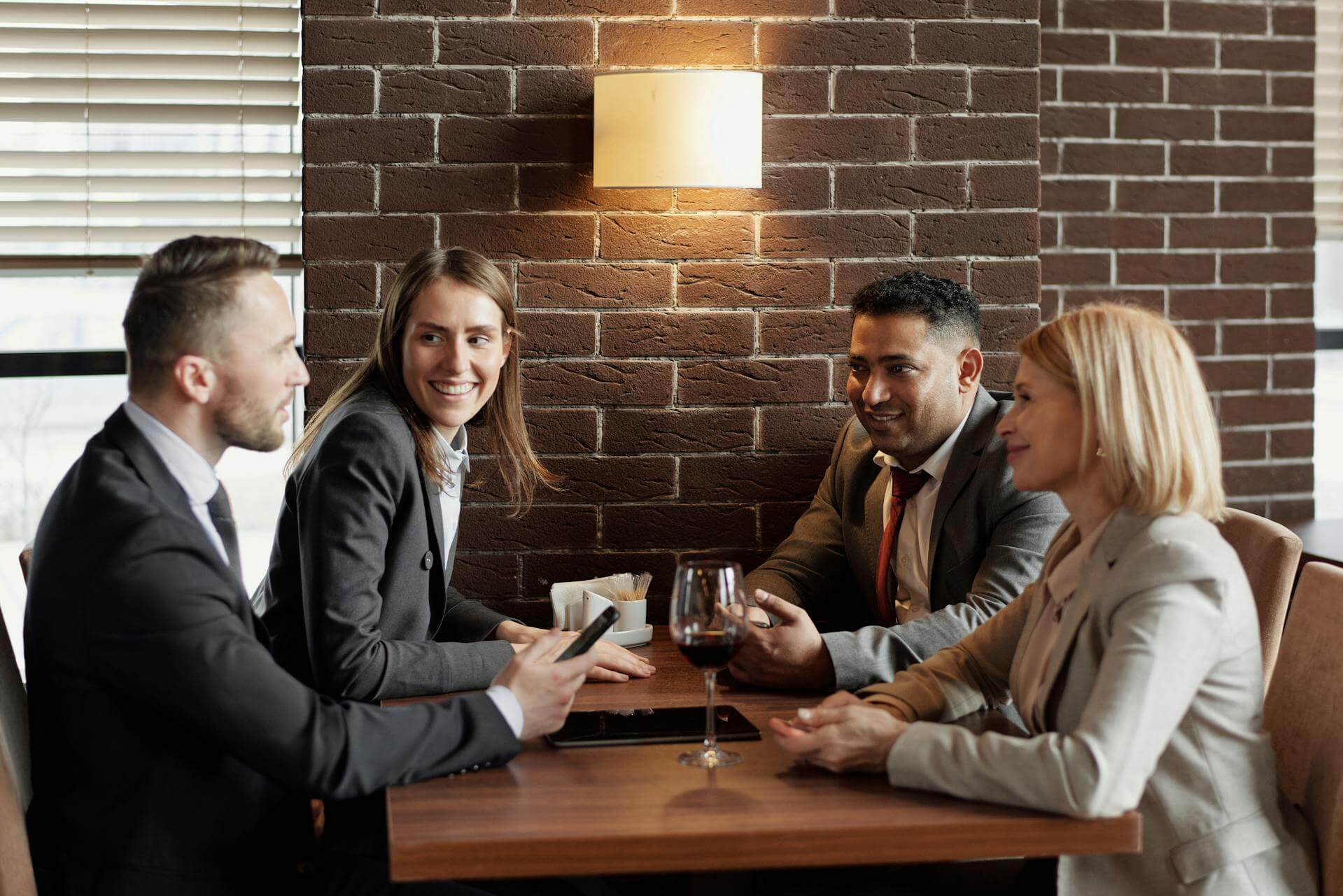 Four professionals in business attire are sitting around a table with drinks, having a conversation in a brick-walled restaurant setting.