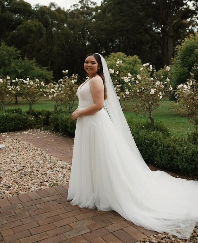 Bride in a white dress and veil stands on a brick path surrounded by greenery and white flowers.