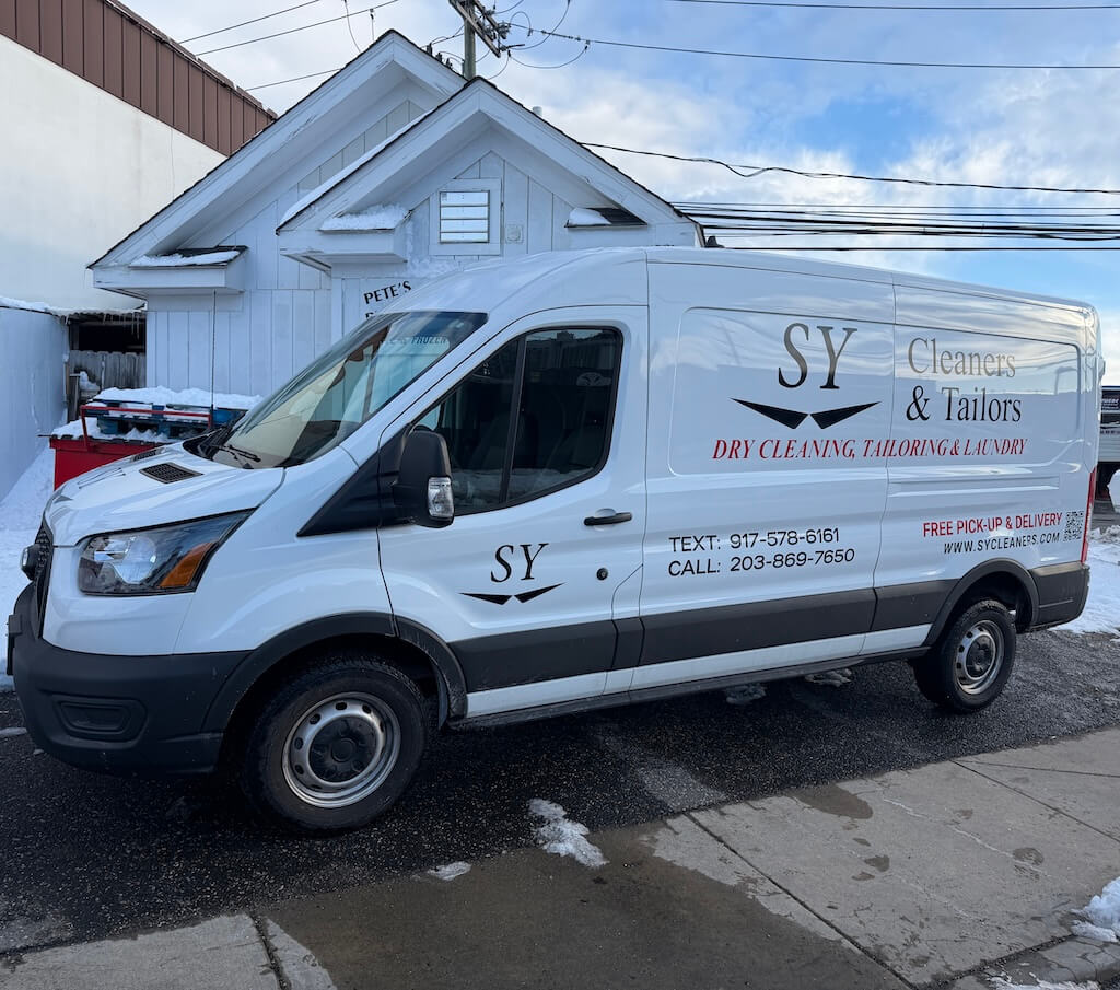 SY CLEANERS DELIVERY TRUCK A white SY Cleaners & Tailors van is parked outside a white building on a snowy day; the van advertises dry cleaning, tailoring, and laundry services with contact numbers.